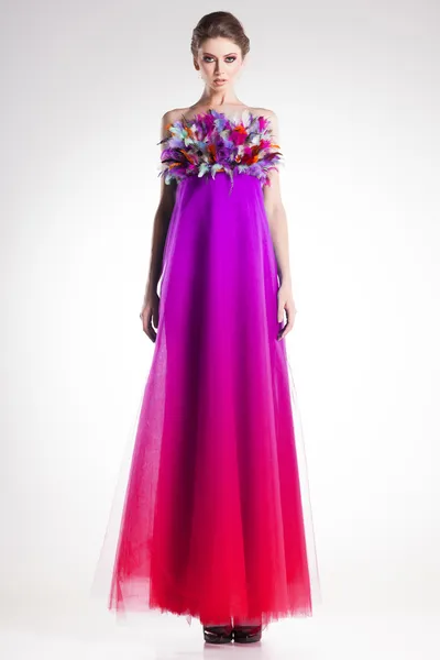 Vacker kvinna modell poserar i långa färgglada klänning med fjädrar i studion — Stockfoto