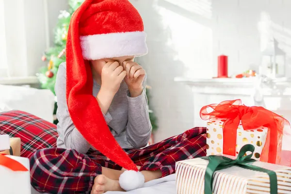 Kırmızı şapkalı üzgün bir çocuk sabahları Noel Baba 'nın hediye kutularının yanında yatağında otururken elleriyle gözlerini ovar ve ağlar. Mutsuz çocuk yeni yıl hediyesiyle hayal kırıklığına uğradı..
