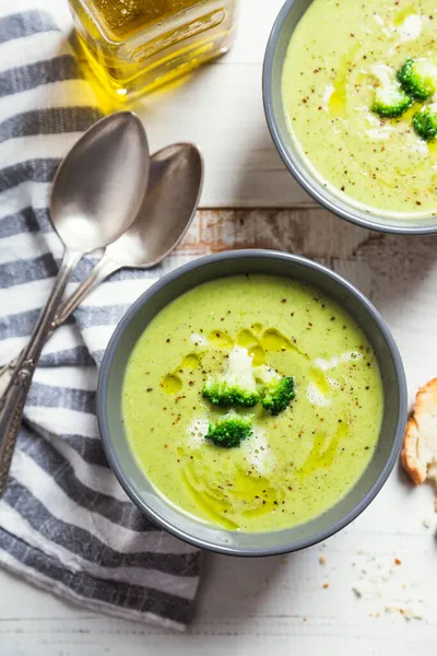 Bir kase brokoli kremalı çorba zeytinyağı, biber ve kızarmış ekmek dilimleri kırsal beyaz ahşap bir masada.