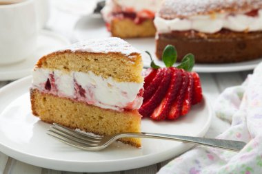 Çilek, reçel ve kremayla dolu Victoria sünger keki. Beyaz ahşap bir masada krema, şeker ve çilekle süslenmiş. 