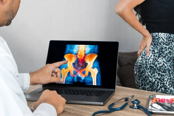 Doctor showing a x-ray of pain in the hips on a laptop. Woman patient in the background. High quality photo