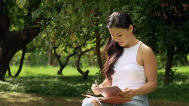 Smiling woman writing in a notebook and laptop. Summer in the park. High quality 4k footage