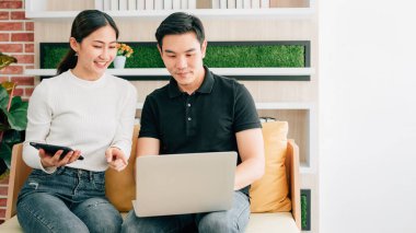 Asyalı erkek, dişinin yanında oturmuş tablet kullanırken gülümsüyor, işaret ediyor, dizüstü bilgisayara birlikte bakıyor. Evde dizüstü bilgisayarla çalışan iş adamları. İyi bir fırsat beraberinde bir sürprizle gelir..