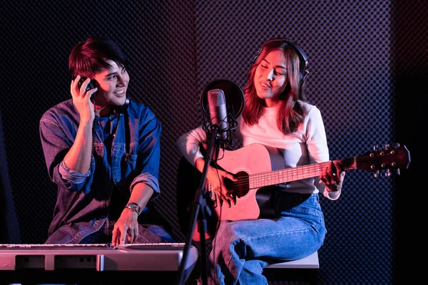 Pretty Asian female singer playing acoustic guitar with male playing electric keyboard. Recording songs by using a studio microphone and pop shield on microphone in blue and red light. Duet session.