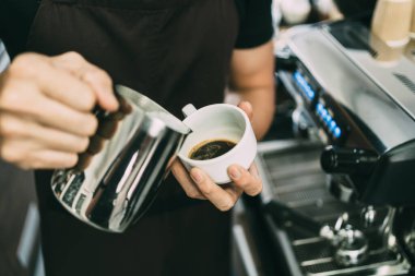 Latte Art yapmak için sıcak espresso sade kahvesine sıcak süt döken genç bir barista görüntüsü..