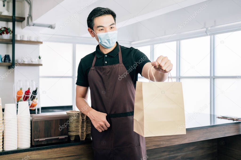 Male barista showing takeaway coffee in the paper bag during Coronavirus outbreak time. A worker inside the cafe counter for online delivery service. Coffee paper bag mockup.