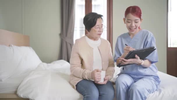 スクラブを装着したアジア系女性看護師は ベッドルームにタブレットを装着したアジア系シニア女性の健康状態を報告しています 介護者が家に来る ホームヘルスケアと介護ホームコンセプト — ストック動画