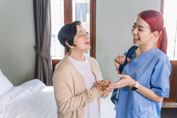 Senior Asian woman talking with Asian woman nurse wearing scrubs in the bedroom. Caregiver visit at home. Home health care and nursing home concept.