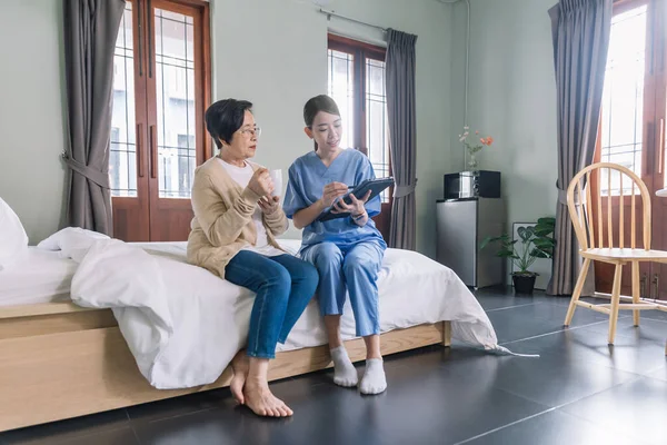 Önlük giyen Asyalı kadın hemşire yatak odasında tabletli yaşlı Asyalı kadının sağlık durumunu rapor ediyor. Bakıcı eve ziyarete geldi. Ev sağlığı ve huzurevi kavramı.