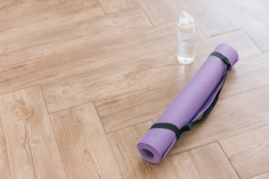 Yuvarlak mor yoga minderi ve temiz plastik su şişesi ahşap bir yüzeyde doğal ışıkla. Evde cinsiyet ayrımı olmayan fitness yogası ve egzersiz aksesuarları veya fotokopi alanı olan spor afişleri. Etkin yaşam biçimi.