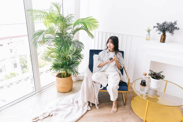 Genç, güzel bir kadın gülümser ve yeşil bir bitkiyle modern yaşam alanının sandalyesine oturur, bir fincan sıcak kahve içerek rahatlar ve rahat bir evde sakin vakit geçirir ve ev ortamının tadını çıkarır..