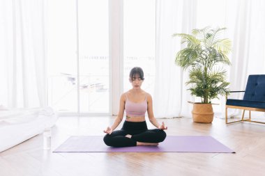 Genç, çekici, sportif Asyalı bir kadın yoga minderinde yoga yapıyor Ardha Padmasana egzersizi yapıyor, yarım Lotus pozu veriyor, evde çalışıyor, spor kıyafetleri giyiyor..
