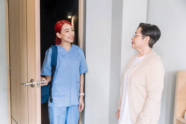 頭蓋骨を着たアジア系女性看護師さんが笑顔で玄関のアジア系の先輩女性を訪ねています ホームヘルスケアと介護ホームコンセプト — ストック写真