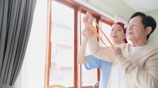 理学療法における高齢患者のための抵抗バンド運動を利用することにより 看護師はアジアの高齢女性とスクラブ演習を身に着けている ホームヘルスケアと介護ホームコンセプト — ストック動画