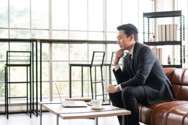 Asian businessman executive manager looking at a laptop computer, thinking about stock and crypto market volatility or meeting an online webinar and waiting for the decision in the office.