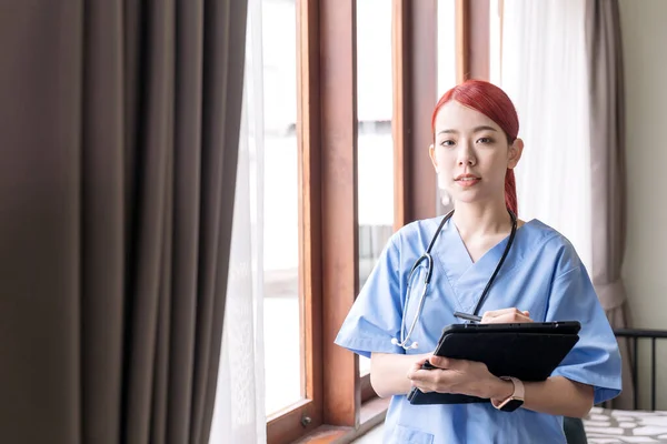 Portrait of Asian woman nurse wearing medical scrubs, stethoscope and holding the tablet in the house of patient. Caregiver visit at home. Home health care and nursing home concept.