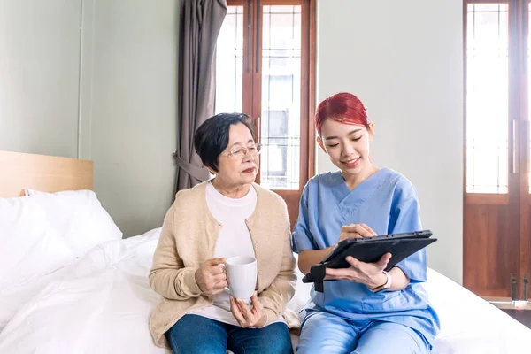 Asian woman nurse wearing scrubs report the health status of Senior Asian woman with digital tablet in the bedroom. Caregiver visit at home. Home health care and nursing home concept.