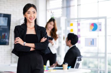 Arka planda buluşan bulanık iş arkadaşlarının önünde siyah ceket giyen gülümseyen Asyalı bir iş kadınının portresi. Kopya alanı ile girişimci liderlik resmi.