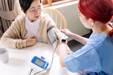 Hemşire, yaşlı Asyalı kadınlarda otomatik tansiyon monitörü kullanarak tansiyonunu ölçüyor. Bakıcı ziyareti. Ev sağlığı ve huzurevi kavramı.