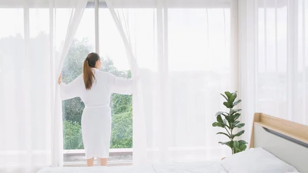Woman wearing bathrobe opens curtains in bedroom and see around the outdoors. Welcome new sunny day, happy positive young woman wakes up and looks in the window at home or hotel feeling optimistic.