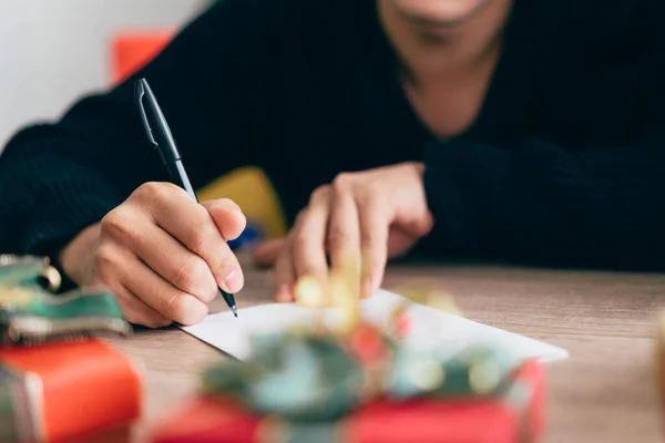 Close up man\'s hand is writing on a blank Christmas postcard with a pen. Couple sitting and writing Christmas card together for sending with surprise gifts at home during Christmas holiday.