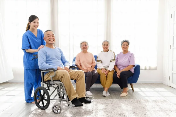 The caregiver therapist stands with an Asian senior sitting in a wheelchair with a group of senior women sitting on a sofa in the background. The nursing home facilitates a support group.