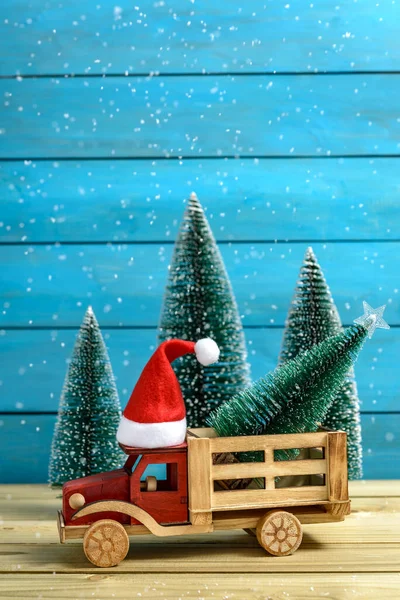 Noel kutlaması konsepti. Noel ağacı, Noel Baba şapkasıyla oyuncak bir kamyonun üstünde, ağaçların yanında. Tahta bir masada kar yağarken..