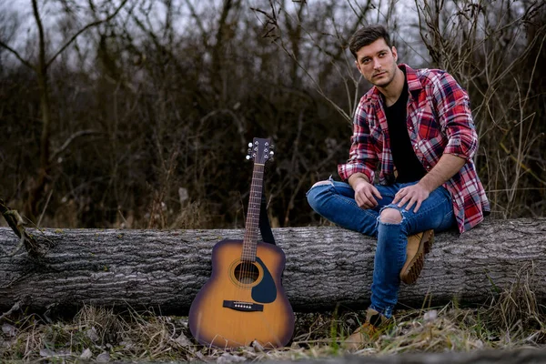 Yakışıklı bir genç adam bir doğa parkında akustik bir gitarın yanında devrilmiş bir ağaçta oturuyor..