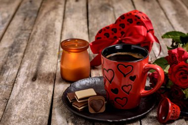 Bir buket kırmızı gül, hediye kutusu, zil, bir fincan kahve ve çikolatalı şekerler ahşap bir masada. Sevgililer Günü için romantik konsept.
