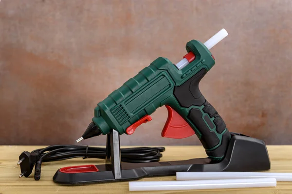 Electric hot glue gun with stand and white glue sticks on wooden background.