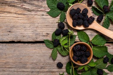 Tahta bir kasede taze böğürtlen ve kırsal bir zeminde yeşil yapraklarla ahşap kaşıkta. Düz yatış, üst görünüm.