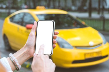 Bir kadın elinde akıllı telefon maketi tutuyor, yakın plan. Şehirdeki bir taksinin arka planında.