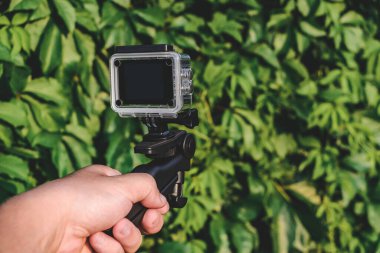 Bir adamın elinde aksiyon kamerası. Aşırı büyümüş yeşil bir çitin arka planında