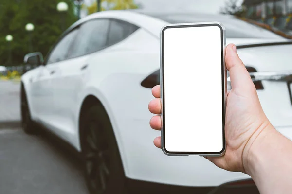 Yakın plan, akıllı telefonun bir erkeğin elinde mokasenti. Şehirde beyaz elektrikli bir arabanın arka planına karşı.