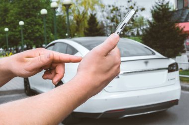 Yakın plan, akıllı telefonun önündeki bir parmak bir adamın ellerindeydi. Şehirde beyaz elektrikli bir arabanın arka planına karşı.