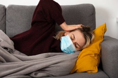 Coronavirus hastası genç bir kadın baş ağrısı ve ateşi var, kanepeye kıvrılmış bir şekilde yatıyor.