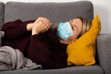 Coronavirus hastası genç bir kadın baş ağrısı ve ateşi var, kanepeye kıvrılmış bir şekilde yatıyor.