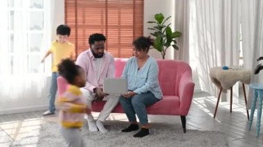 4K Happy parents couple sit on couch using laptop with children running around