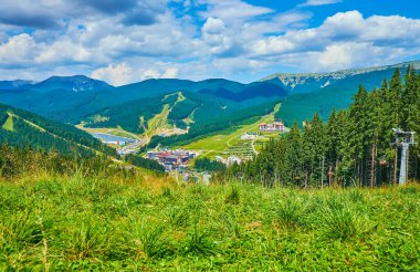 Bukovel Dağı tepesi, yemyeşil dağ çayırının, Bukovel tatil köyünün, teleferiklerin, diğer dağ yamaçlarının ve ormanların, Karpatların, Ukrayna 'nın manzarasını açar.