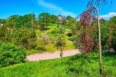 Kyiv Botanik Bahçesi 'nin tepelik manzarası yemyeşil, çiçek açan ağaçlar ve çalılar ve arka planda Aziz Jonas Trinity Manastırı' nın kubbesi, Ukrayna