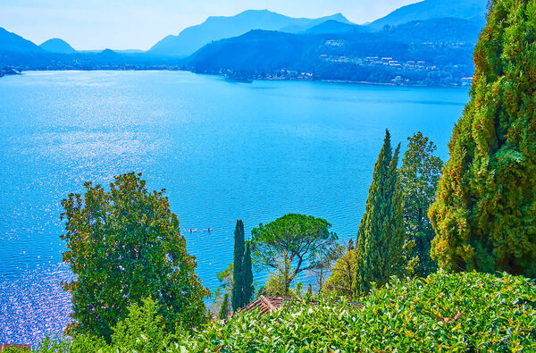 The scenic Alpine landscape and bright blue rippled surface of Lake Lugano behind the lush green trees and shrubs of Scherrer Park, Morcote, Switzerland