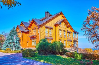 MEZHYHIRYA, UKRAINE - OCT 26, 2021: The green lawn, conifer trees and bushes in autumn park around the wooden Honka club house, on Oct 26 in Mezhyhirya