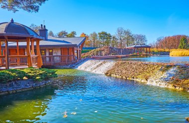 Küçük şelalesi, ahşap çardağı ve arka planda verimli bir sonbahar parkı olan güzel göl, Mezhyhirya, Ukrayna