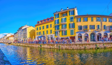 MILAN, ITALY - APRIL 9, 2022: The crowded embankment of Naviglio Grande Canal with old colorful houses and restaurants behind pedestrian alley, on April 9 in Milan