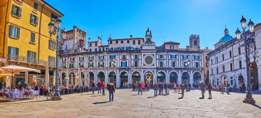 BRESCIA, ITALY - APRIL 10, 2022: Panoramic view of historic building with arcades and Torre dell'Orologio (Clock Tower) with astronomical clock and bright sunshine in the background, on April 10 in Brescia