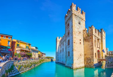 SIRMIONE, ITALY - APRIL 10, 2022: The main historic landmark of Sirmione - Castello Scaligero with moat and colored houses of Via Dante street around it, on April 10 in Sirmione