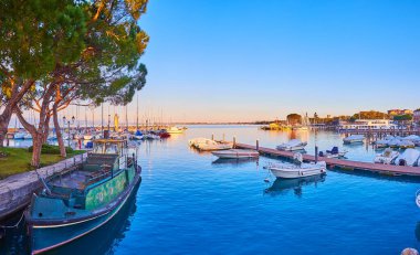 The sunset over the port with a military boat, yachts and fishing boats moored at the banks and shipyards, Desenzano del Garda, Lake Garda, Italy