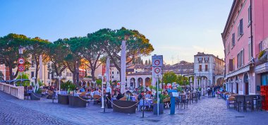 DESENZANO DEL GARDA, ITALY - APRIL 10, 2022: The evening panorama of the Old Port with outdoor restaurants and tall pines, on April 10 in Desenzano del Garda