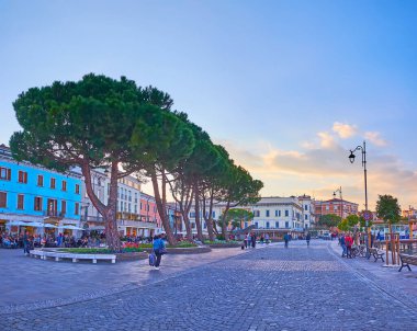 DESENZANO DEL GARDA, ITALY - APRIL 10, 2022: The evening embankment of Lake Garda with old houses, restaurants, spread pines and flower beds, Lungolago Cesare Battisti, on April 10 in Desenzano del Garda