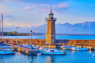 Historic stone lighthouse on Diga Foranea breakwater with yachts and boats in port and the Garda Prealps in the background at foggy sunset, Desenzano del Garda, Lake Garda, Italy